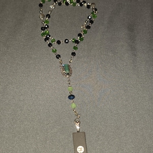 May include: A rosary necklace with a silver chain and a mix of green, black, and clear beads. The pendant features a rectangular, dark gray charm with text and a circular logo. The beads are arranged in a traditional rosary design.
