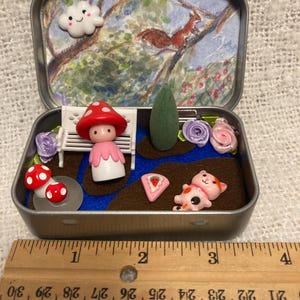 May include: A miniature garden scene inside a small, open metal tin. The scene includes a painted backdrop of a squirrel in a tree, a mushroom-topped figure, a white bench, and various miniature elements like flowers and a cat. The tin is approximately 4 inches long.