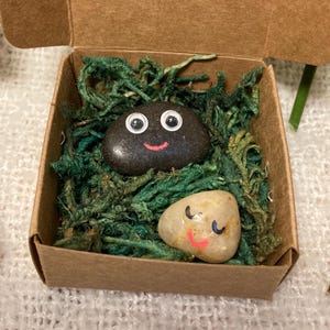 May include: Two painted rocks with smiling faces rest in a small cardboard box filled with green moss. One rock is dark gray with white eyes and a red smile. The other is light beige with closed eyes and a pink smile. A unique gift idea.