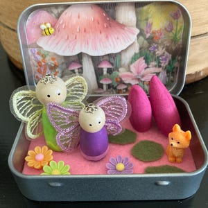 May include: A metal tin containing two wooden peg dolls with sheer wings, one in green and one in purple. The scene includes a small orange fox figurine, two pink cone-shaped items, felt circles, and plastic flowers, set against a mushroom backdrop.