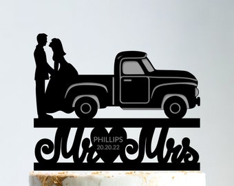 May include: Black silhouette of a couple standing in the back of a pickup truck with the words "Mr & Mrs PHILLIPS 20.20.22" on a heart-shaped background. The topper is on top of a two-tiered wedding cake.