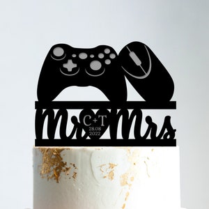 May include: Black and white cake topper featuring a video game controller and a computer mouse. The topper also includes the text "Mr & Mrs" with a heart and the date "28.08.2022".