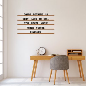 May include: A wooden letter board with the quote "DOING NOTHING IS VERY HARD TO DO... YOU NEVER KNOW WHEN YOU'RE FINISHED." The board is hanging on a white wall above a wooden desk with a chair.