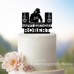 Op de afbeelding: Een zwarte taarttopper met een DJ-silhouet, koptelefoon, luidsprekers en het nummer 20. De tekst luidt "HAPPY BIRTHDAY ROBERT". De topper staat op een witte taart, versierd met bloemen.