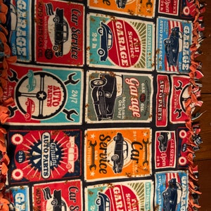 Op de afbeelding: Een zwarte fleece deken met een herhaald patroon van vintage autoservicetegels in rood, oranje, blauw en blauwgroen. De tegels tonen klassieke auto's, moersleutels en de woorden "Garage" en "Car Service". De deken heeft een oranje franje.