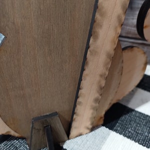 Op de afbeelding: Een houten uitsnijding van een hart met een decoratieve rand, staande op een zwarte houten standaard. Het hart is lichtbruin van kleur en heeft een donkerbruine rand.