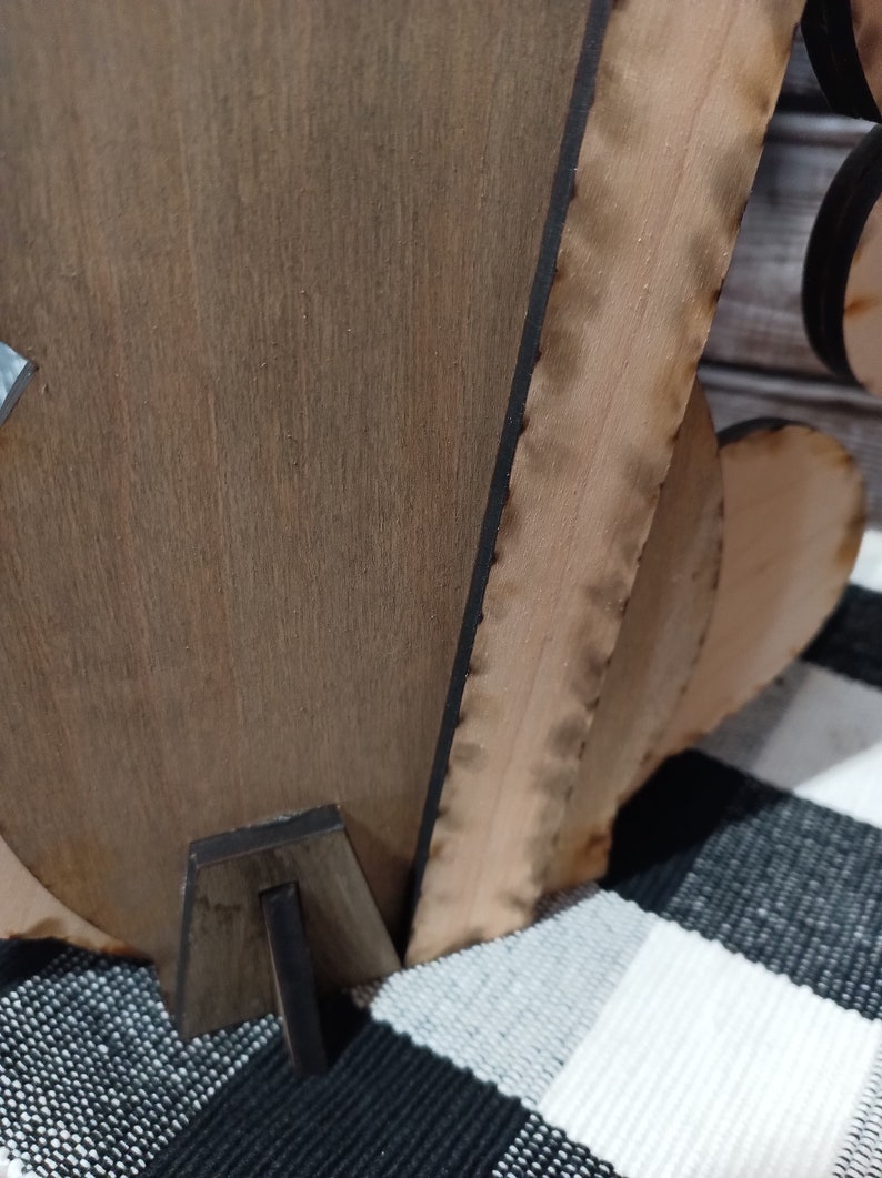 Op de afbeelding: Een houten uitsnijding van een hart met een decoratieve rand, staande op een zwarte houten standaard. Het hart is lichtbruin van kleur en heeft een donkerbruine rand.