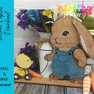 May include: A brown bunny wearing blue overalls and a white shirt. The bunny is holding a carrot in its right hand. The bunny is standing on a wooden surface with a green background. The Digital Makery text is visible in the top right corner of the image.