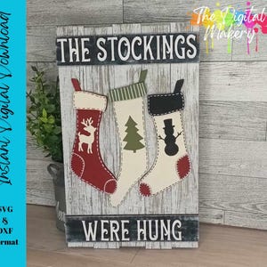 May include: A wooden sign with the text "The Stockings Were Hung" and three decorative stockings. The stockings are red, green, and white with a snowman, a tree, and a reindeer design.