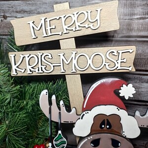 Op de afbeelding: Een houten kerstbord met de woorden "Merry Kris-Moose" in witte letters. Een bruine eland met een rode kerstmanmuts en witte bontkraag houdt groene en rode ornamenten vast.