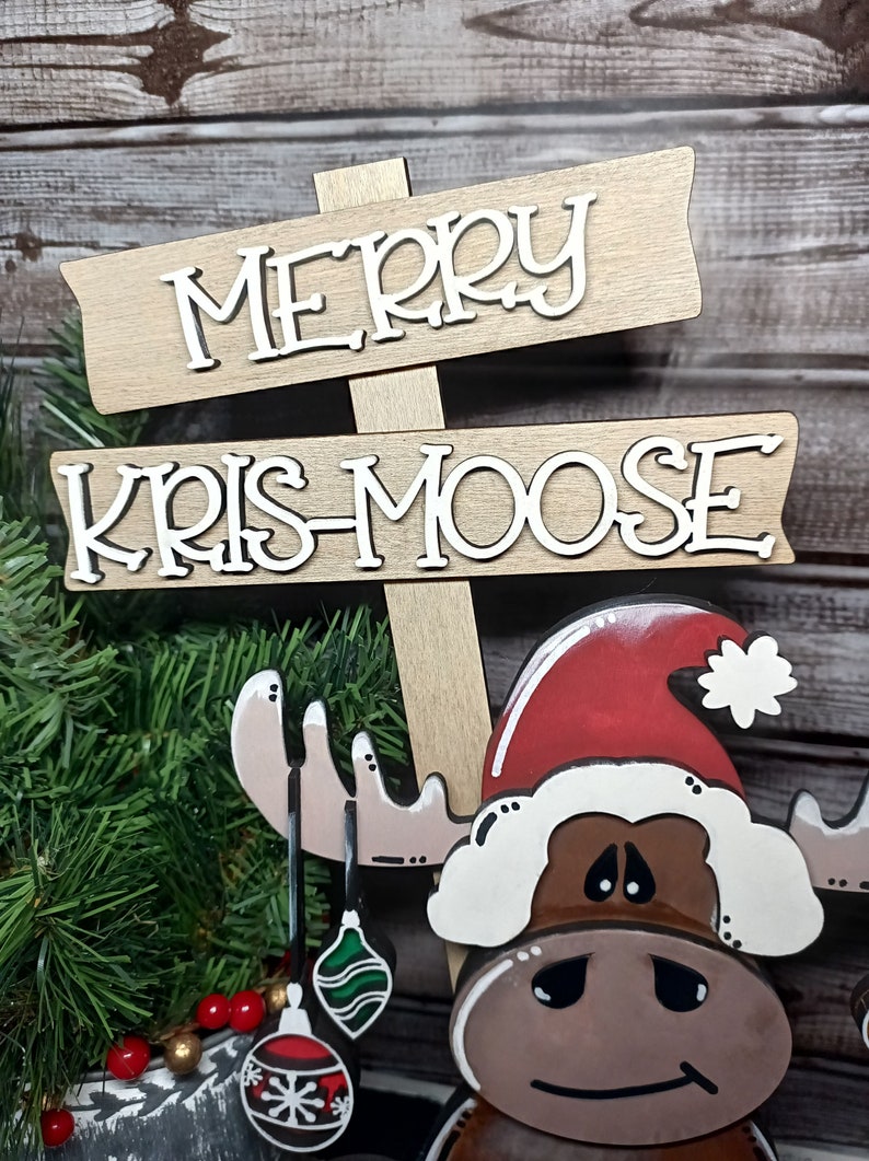 Op de afbeelding: Een houten kerstbord met de woorden "Merry Kris-Moose" in witte letters. Een bruine eland met een rode kerstmanmuts en witte bontkraag houdt groene en rode ornamenten vast.