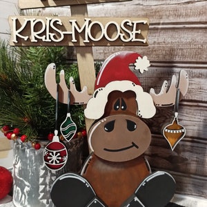 Op de afbeelding: Houten kerst-elchfiguur met een kerstmanmuts en kerstballen. De elch is bruin met zwarte hoeven en een witte baard. Het bord erboven staat "Merry Kris-Moose".