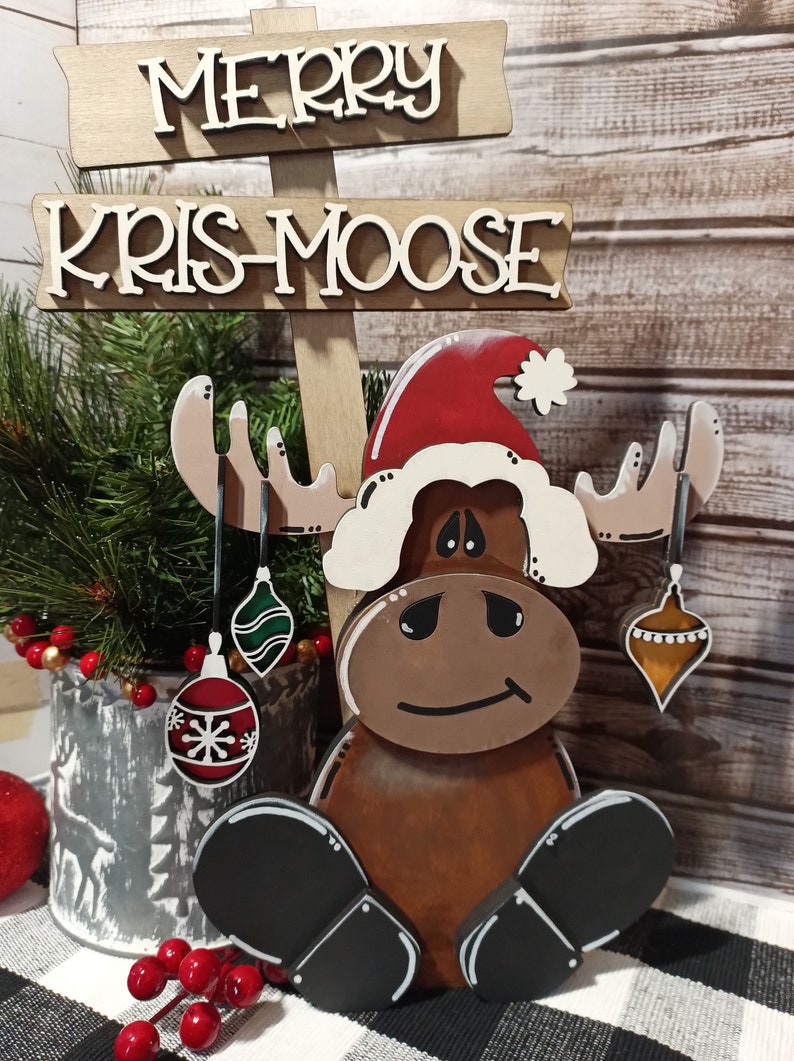 Op de afbeelding: Houten kerst-elchfiguur met een kerstmanmuts en kerstballen. De elch is bruin met zwarte hoeven en een witte baard. Het bord erboven staat "Merry Kris-Moose".