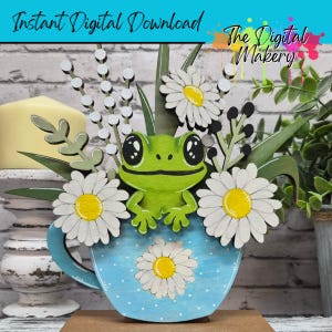 Könnte beinhalten: Eine skurrile Holzfroschfigur in einer blauen Teetasse mit weißen Gänseblümchen und Grün. Die Teetasse hat ein Gänseblümchen-Design. Die Worte "Instant Digital Download" und "The Digital Makery" sind sichtbar.