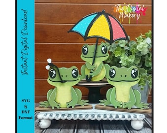 Peut inclure: Trois découpes de grenouilles vertes avec de grands yeux, dont une tenant un parapluie coloré. Les grenouilles sont sur un présentoir décoratif blanc. Le texte "Instant Digital Download" et "SVG & DXF Format" sont visibles.