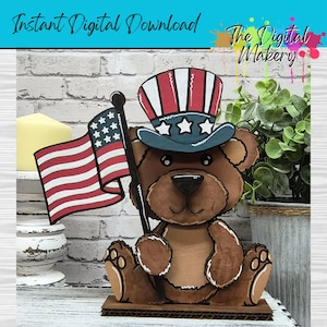 May include: A brown teddy bear figure wearing a patriotic hat and holding an American flag. The bear is on a cardboard base. The image includes the text "Instant Digital Download" and "The Digital Makery".