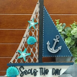K&ouml;nnte beinhalten: Holz-Segelboot-Dekoration mit blauem Segel und Anker sowie einem hellen Segel mit Gittermuster. Verziert mit t&uuml;rkisfarbenen Seesternen und Muschelakzenten. Auf der Basis steht "Seas The Day".