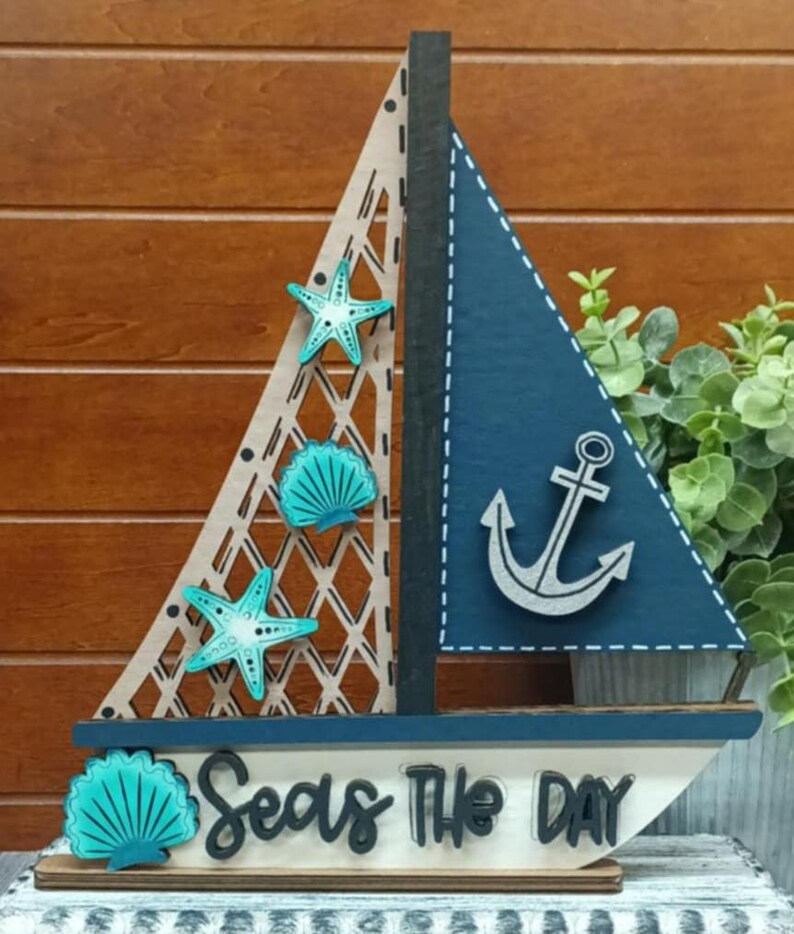 K&ouml;nnte beinhalten: Holz-Segelboot-Dekoration mit blauem Segel und Anker sowie einem hellen Segel mit Gittermuster. Verziert mit t&uuml;rkisfarbenen Seesternen und Muschelakzenten. Auf der Basis steht "Seas The Day".