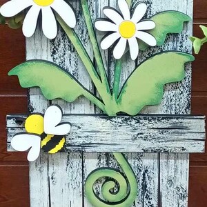May include: A white wooden sign with a distressed finish features a painted design of three white daisies with yellow centers and green leaves. A yellow and black bee is perched on a wooden plank with a white background. The sign has a green swirl at the bottom.