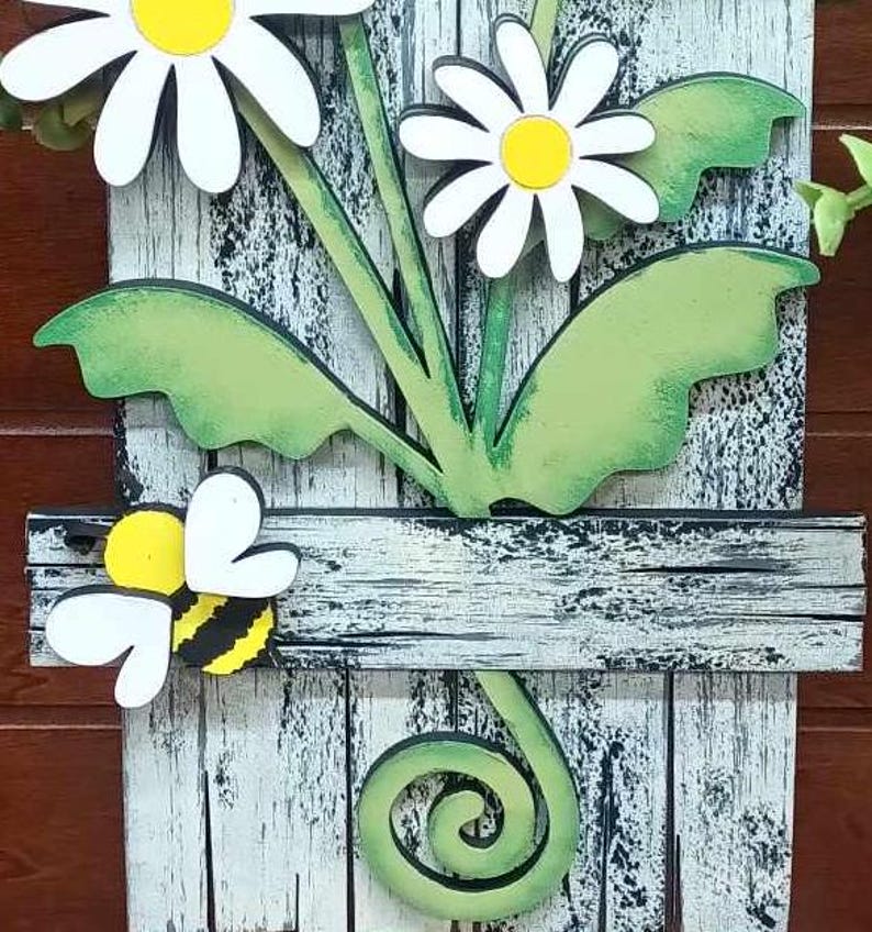 May include: A white wooden sign with a distressed finish features a painted design of three white daisies with yellow centers and green leaves. A yellow and black bee is perched on a wooden plank with a white background. The sign has a green swirl at the bottom.