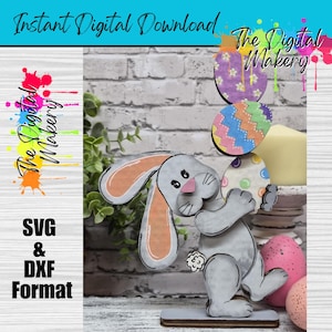Könnte beinhalten: Eine Holzfigur eines Osterhasen, der dekorierte Ostereier hält. Der Hase ist grau mit orangefarbenen Ohren und einem weißen Schwanz. Das Bild enthält den Text "Instant Digital Download" und "SVG & DXX Format".