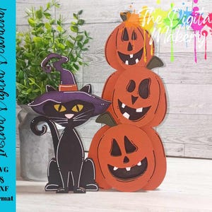 Puede incluir: Un gato negro con un sombrero de bruja se sienta frente a una pila de tres calabazas naranjas con caras talladas. Las calabazas están apiladas una encima de la otra, con la calabaza más grande en la parte inferior. El gato está mirando a la cámara.