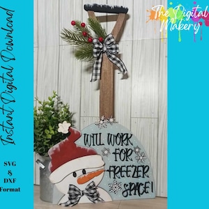 May include: A wooden shovel-shaped sign with a snowman design and the text "WILL WORK FOR FREEZER SPACE!". The snowman wears a red hat and a black and white checkered bow. The shovel handle is decorated with greenery and a bow.