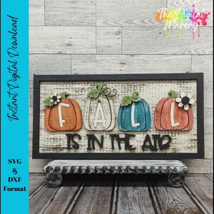 以下が含まれることがあります： 黒いフレームの長方形の木製サインで、「FALL IS IN THE AIR」のフレーズとオレンジ、白、ティール色のカボチャの切り抜きが4つあります。このサインは、装飾的で使い古された白いスタンドに表示されています。