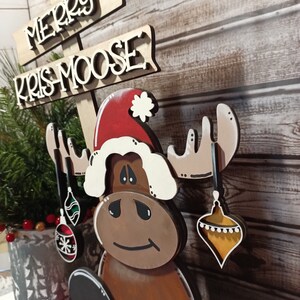 Op de afbeelding: Een houten elandbeeldje met een kerstmanmuts en kerstballen. De eland zit en heeft een bruin lichaam, witte geweien en een bruin gezicht. Het bord boven de eland staat "Merry Kris Moose".