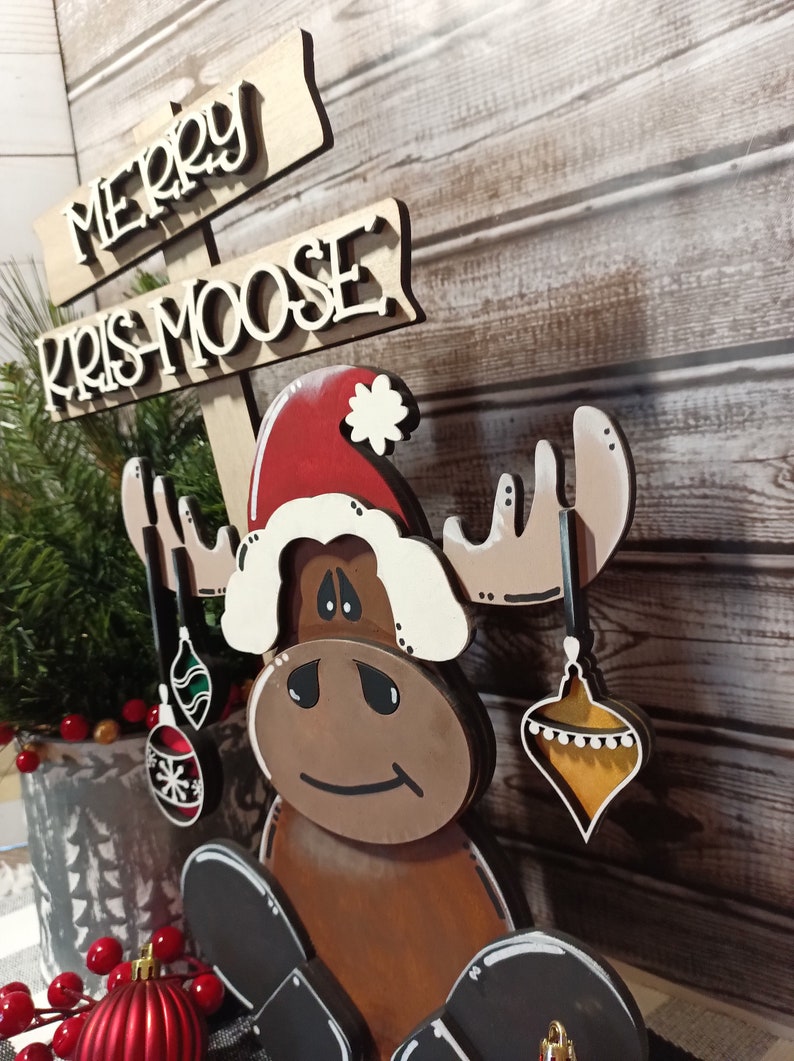 Op de afbeelding: Een houten elandbeeldje met een kerstmanmuts en kerstballen. De eland zit en heeft een bruin lichaam, witte geweien en een bruin gezicht. Het bord boven de eland staat "Merry Kris Moose".