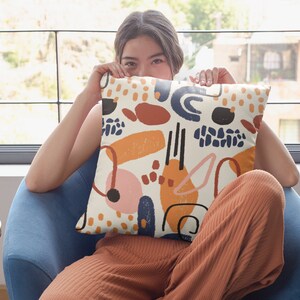 K&ouml;nnte beinhalten: Ein quadratisches Dekokissen mit cremefarbenem Hintergrund und einem verstreuten Muster aus abstrakten Formen in Marineblau, gebranntem Orange, Senfgelb und zartem Rosa. Das Design zeigt verschiedene Pinselstriche, Punkte und geschwungene Linien.