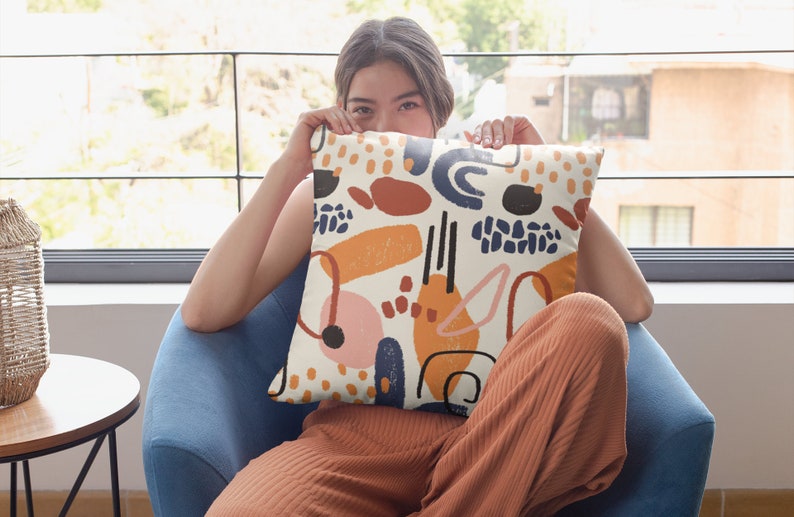 K&ouml;nnte beinhalten: Ein quadratisches Dekokissen mit cremefarbenem Hintergrund und einem verstreuten Muster aus abstrakten Formen in Marineblau, gebranntem Orange, Senfgelb und zartem Rosa. Das Design zeigt verschiedene Pinselstriche, Punkte und geschwungene Linien.