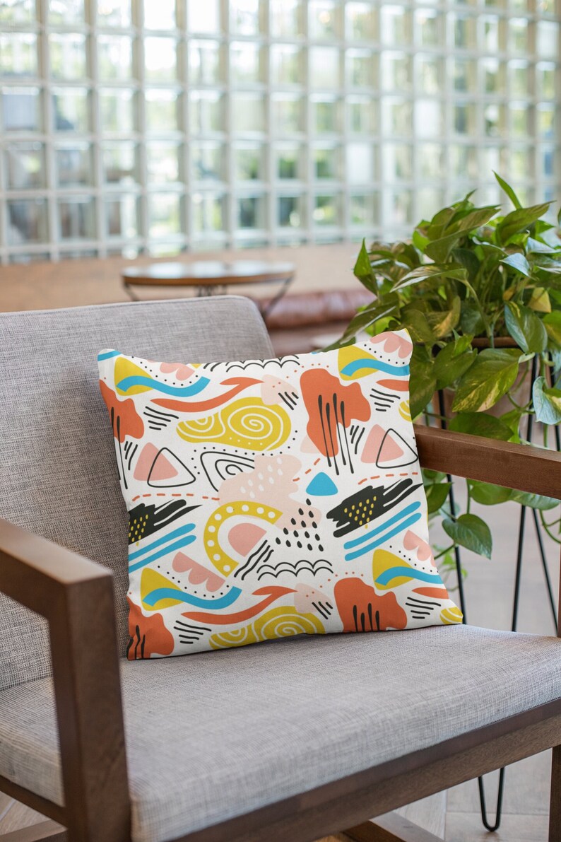 K&ouml;nnte beinhalten: Ein quadratisches Dekokissen mit wei&szlig;em Hintergrund und einem sich wiederholenden Muster aus abstrakten Formen in Gelb, Orange, Hellblau, Rosa und Schwarz. Das Design umfasst Wellenlinien, gef&uuml;llte Fl&auml;chen, Dreiecke, Punkte und Striche.