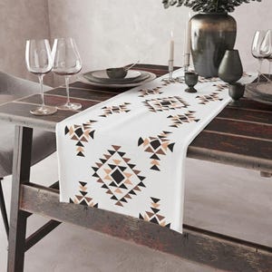 May include: A white table runner featuring a repeating geometric pattern of black and tan triangles and diamonds. The runner is laid across a dark wooden table set with clear wine glasses, a large dark vase, and small black decorative bowls.