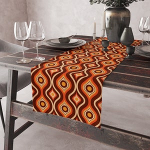 Peut inclure: Un chemin de table brun, orange et crème avec un motif géométrique. Le chemin de table est sur une table en bois avec deux verres à vin et un vase.