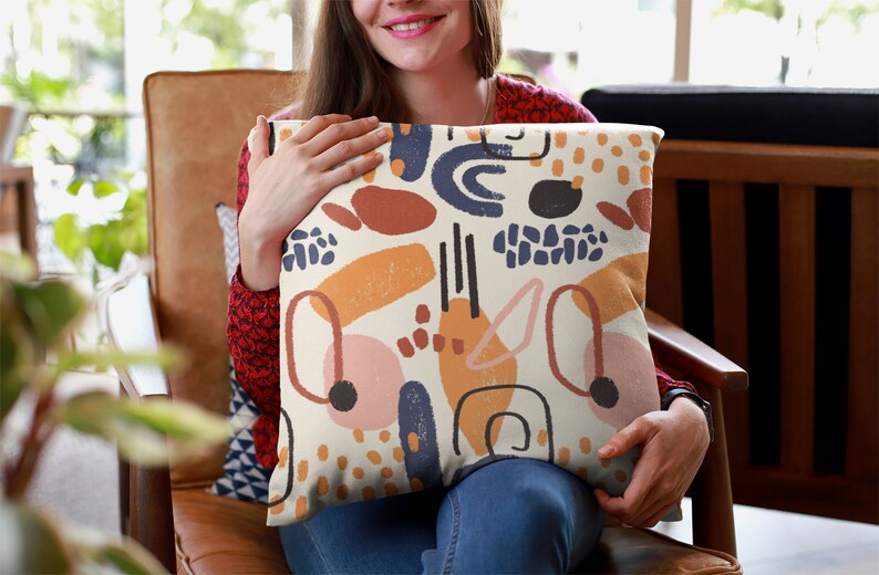 K&ouml;nnte beinhalten: Ein quadratisches Zierkissen mit cremefarbenem Hintergrund und einem wiederkehrenden Muster aus abstrakten Formen in Marineblau, Terrakotta, Senfgelb und zartem Rosa. Das Design zeigt verschiedene handgezeichnete Linien, Punkte und Fl&auml;chen.