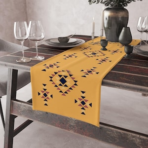 May include: A yellow table runner with a geometric pattern in shades of blue, red, and black. The runner is on a wooden table with two wine glasses and a vase.
