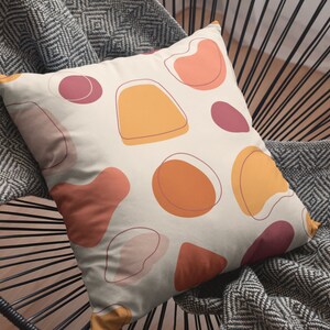 K&ouml;nnte beinhalten: Ein quadratisches Dekokissen mit cremefarbenem Hintergrund und einem wiederkehrenden Muster aus abstrakten organischen Formen in den Farben gebranntes Orange, Senfgelb und Dunkelrot, akzentuiert durch d&uuml;nne, dunkle Umrandungen.