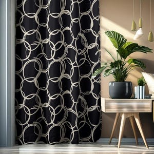 May include: Black curtain with a repeating pattern of off-white overlapping circles. The curtain hangs next to a window and a small wooden table with a potted plant. The room has a neutral color scheme.