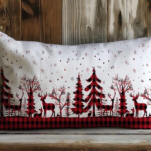 May include: A white lumbar pillow with a red and black buffalo plaid border. The pillow features a winter scene with black silhouettes of trees, deer, and snowflakes on a white background.