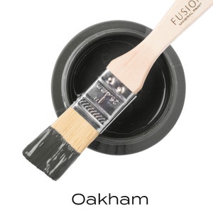 Può includere: Un piccolo barattolo di Fusion Mineral Paint nel colore Oakham. La vernice è di colore grigio scuro. Un pennello con vernice grigia sulle setole è appoggiato sul barattolo.