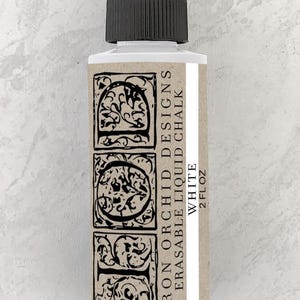 May include: A clear glass bottle with a black cap, containing white erasable liquid chalk. The bottle label features the text "Iron Orchid Designs Erasable Liquid Chalk White 2 fl oz".