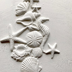 May include: White painted seashells, starfish, and a seahorse arranged in a vertical stack on a white textured background.