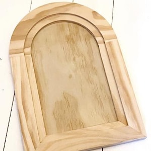 May include: A light-colored, unfinished wooden arched frame. The frame has a rectangular base with an arched top, featuring a recessed panel. The wood grain is visible, and the frame is set against a white background.