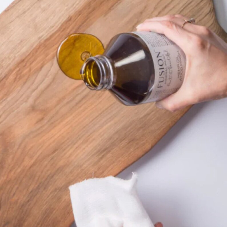 May include: A hand pouring a dark brown liquid from a bottle labeled "FUSION" onto a wooden cutting board. The liquid is spreading out on the board, creating a wet, shiny surface.