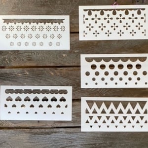 May include: Five white stencils with different patterns. The patterns include a scalloped edge with small flowers, a scalloped edge with polka dots, a scalloped edge with hearts, a scalloped edge with a geometric pattern, and a scalloped edge with a floral pattern.