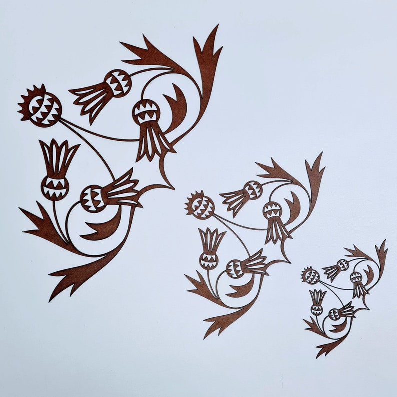 May include: A brown paper cut-out design of a stylized thistle flower with multiple repeating patterns. The design is symmetrical and features a series of overlapping thistle flowers with intricate details.