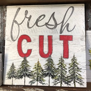 May include: A white wooden sign with the words "fresh cut" in black and red lettering. The sign has a rustic look and features a row of five green pine trees at the bottom.