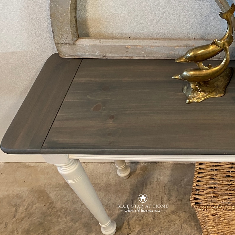 May include: A dark gray wooden console table with white painted legs. The table top has a distressed finish and is decorated with a gold metal dolphin sculpture. The table is sitting on a concrete floor.