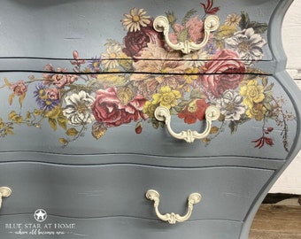 Puede incluir: Una cómoda antigua pintada de gris con dos cajones y calcomanías florales ornamentadas. La cómoda tiene tiradores de porcelana blanca y un borde inferior festoneado.